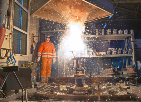 Photo shows a workshop where yellow sparks fly during a chemical reaction. A man in work clothes and protective goggles is standing next to it at some distance.