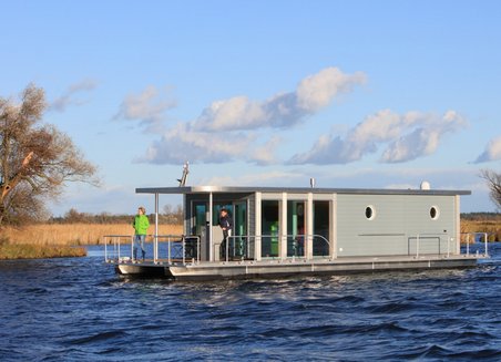 A houseboat floats on the water. It is grey, two people are standing on the boat terrace at the front end. In the background you can see fields, trees and blue sky with clouds.