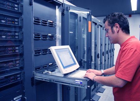 A man is working standing at a laptop. He is in a room in a data center.