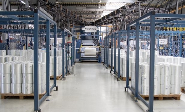 In a factory building, several rolls of material are stored on top of each other to the right and left of an aisle.