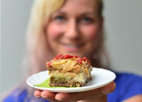Owner Kirstin Knufmann can be seen blurred in the background. She is holding a piece of cake on a small plate towards the camera.